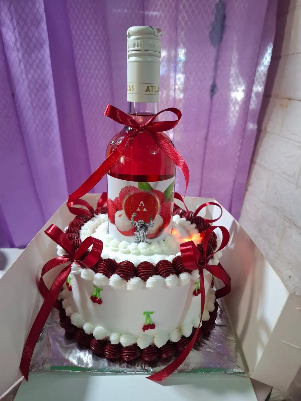 A white frosted cake decorated with red ribbons and cherries, topped with a wine bottle.