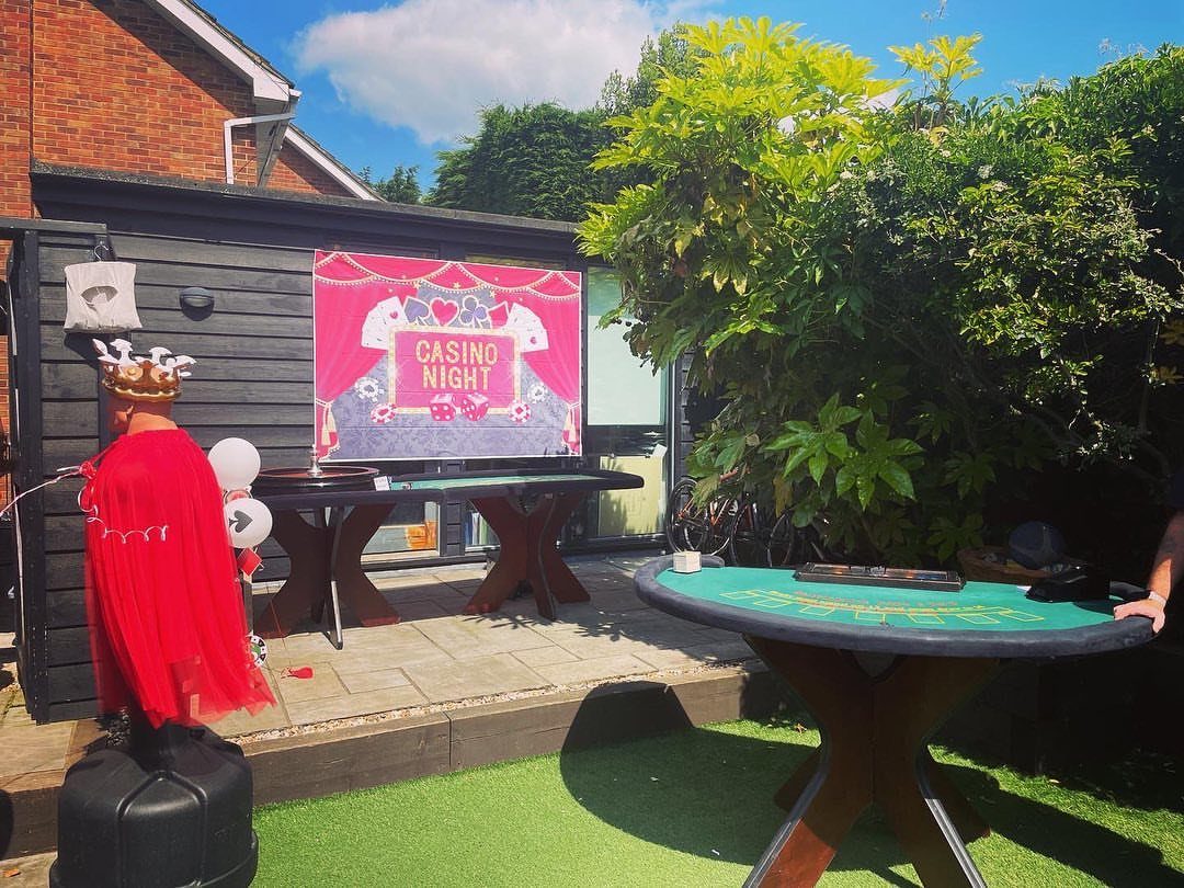 Casino night setup outdoors with a large pink sign, roulette table, and a person in a red cape.