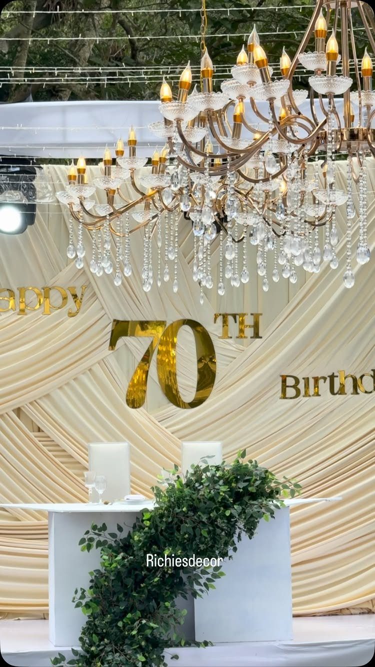 Gold 70th birthday sign against cream draped fabric with a crystal chandelier and greenery.