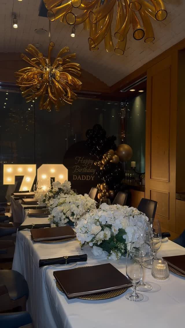 Decorated dining table with white flowers, wine glasses, and large light-up numbers seventy in the background.