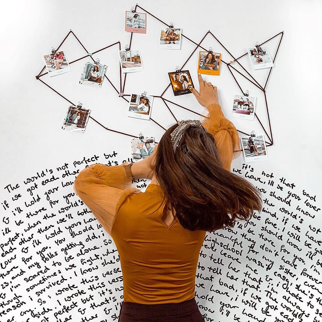 Woman hanging polaroid photos on geometric wire against a wall covered in handwritten lyrics.
