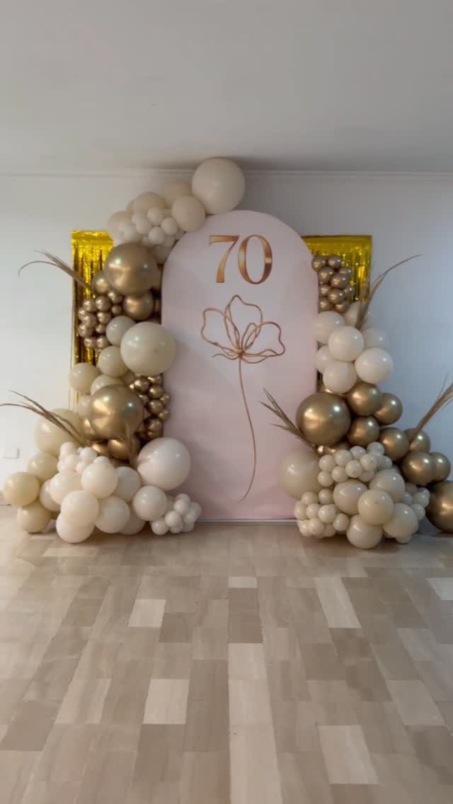 A pink backdrop with the number 70 surrounded by white and gold balloon arches.