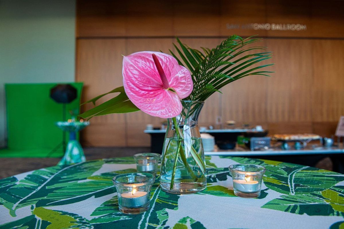 Tropical table centerpiece with pink anthurium, palm leaves, and candle holders on a leaf-print tablecloth.