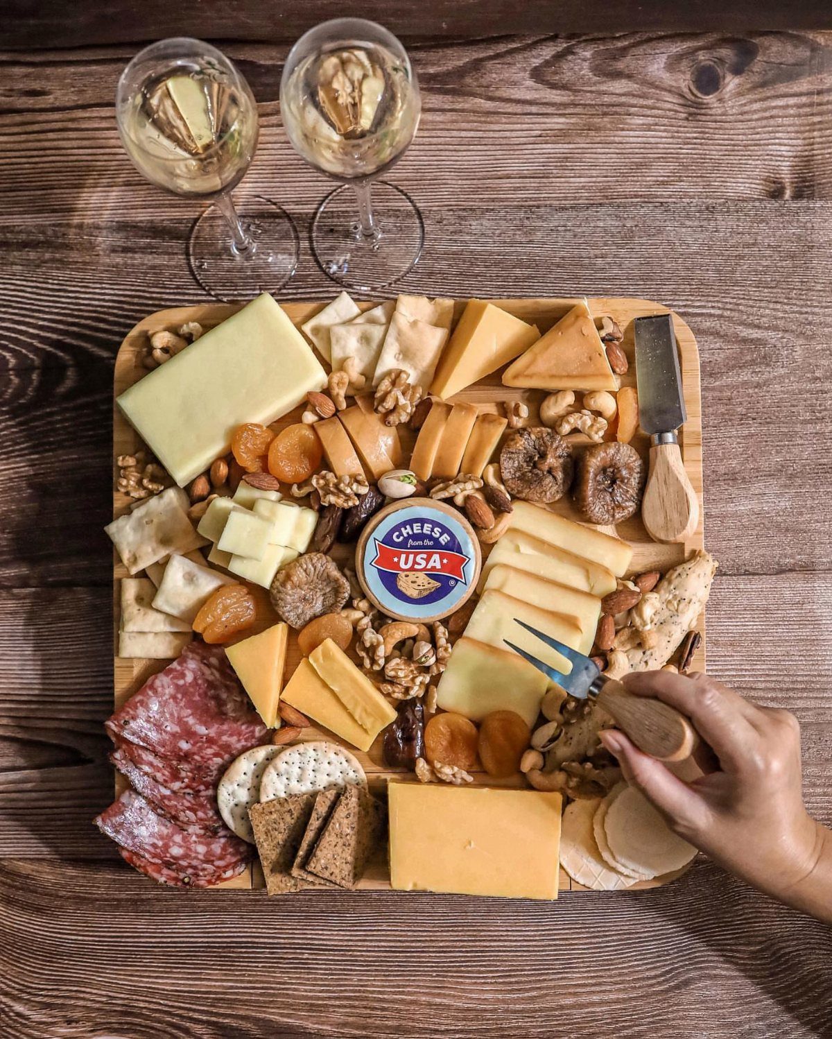 Overhead shot of a wooden cheese and charcuterie board with two glasses of white wine on a rustic table.