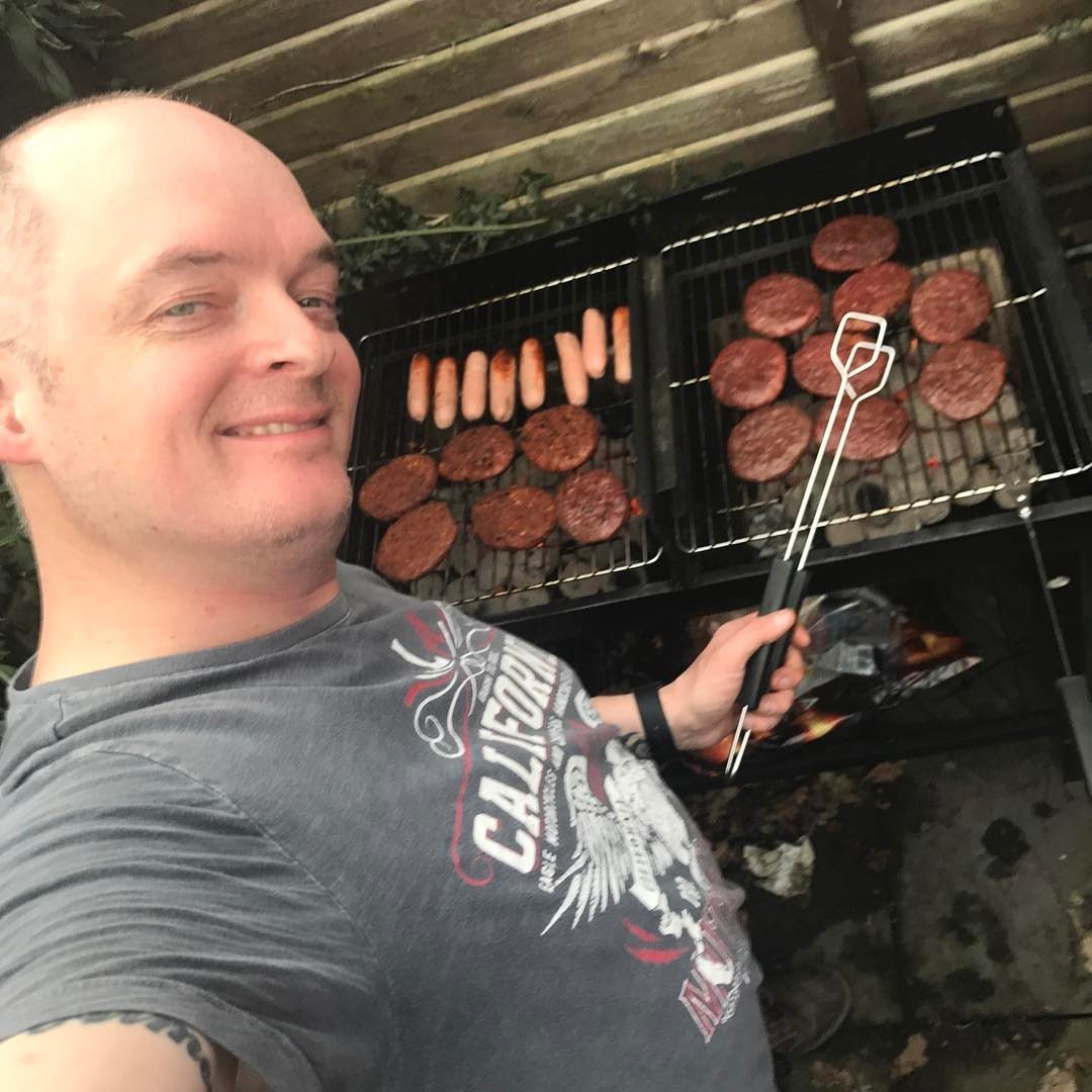 Smiling man taking a selfie holding tongs in front of an outdoor smoker full of cooking burgers and sausages.