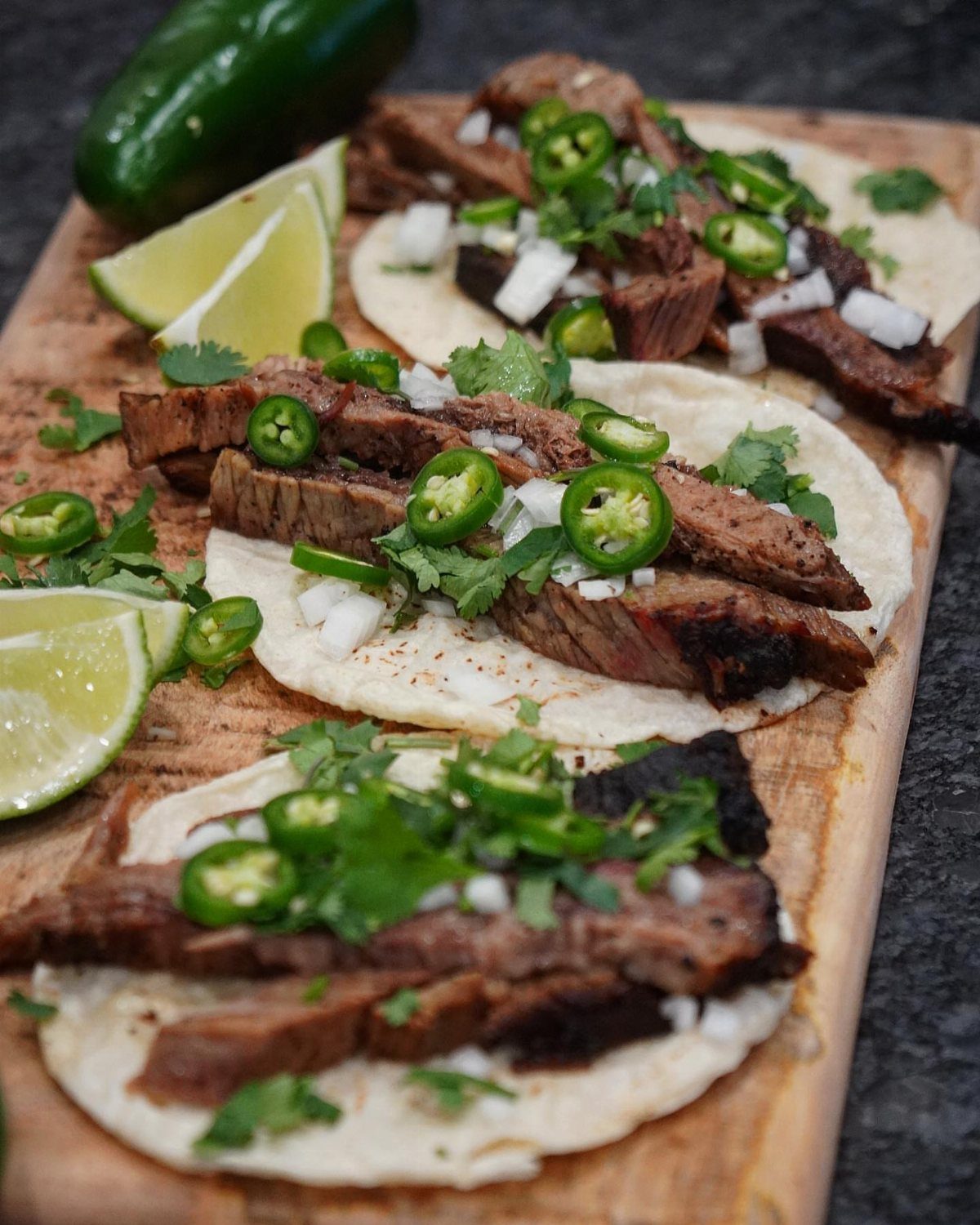 Three small brisket tacos topped with onions, cilantro, and jalapenos on a wooden board with lime wedges.