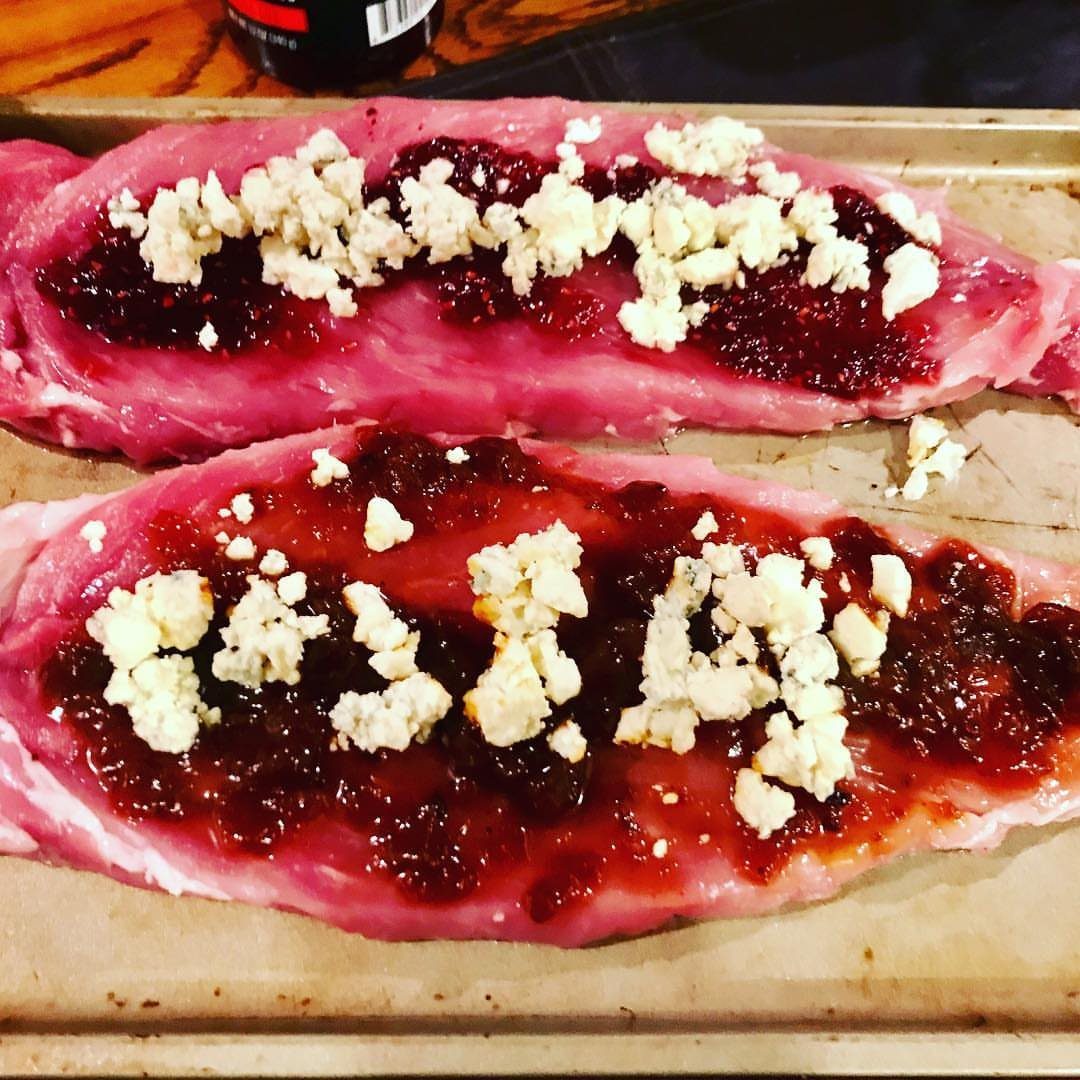 Raw stuffed pork tenderloins topped with cranberry jam and crumbled blue cheese on parchment paper.