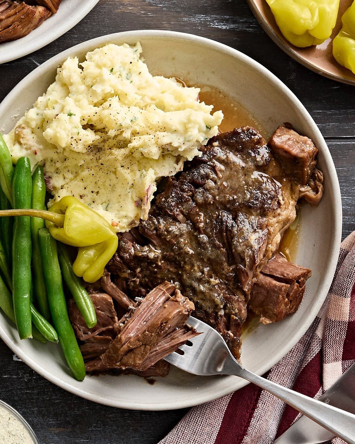 Bowl of sliced pot roast with gravy, mashed potatoes, green beans, and peppers on dark wood table.