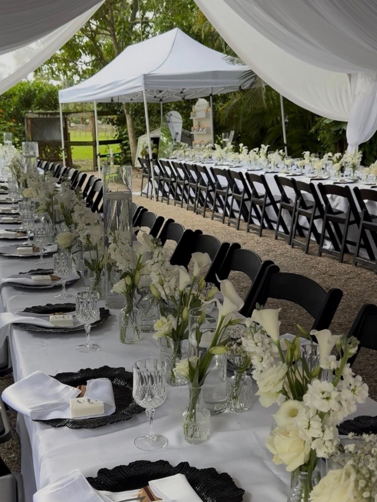 Long outdoor banquet tables set with white linens, black chairs, and white calla lily centerpieces under a draped canopy.