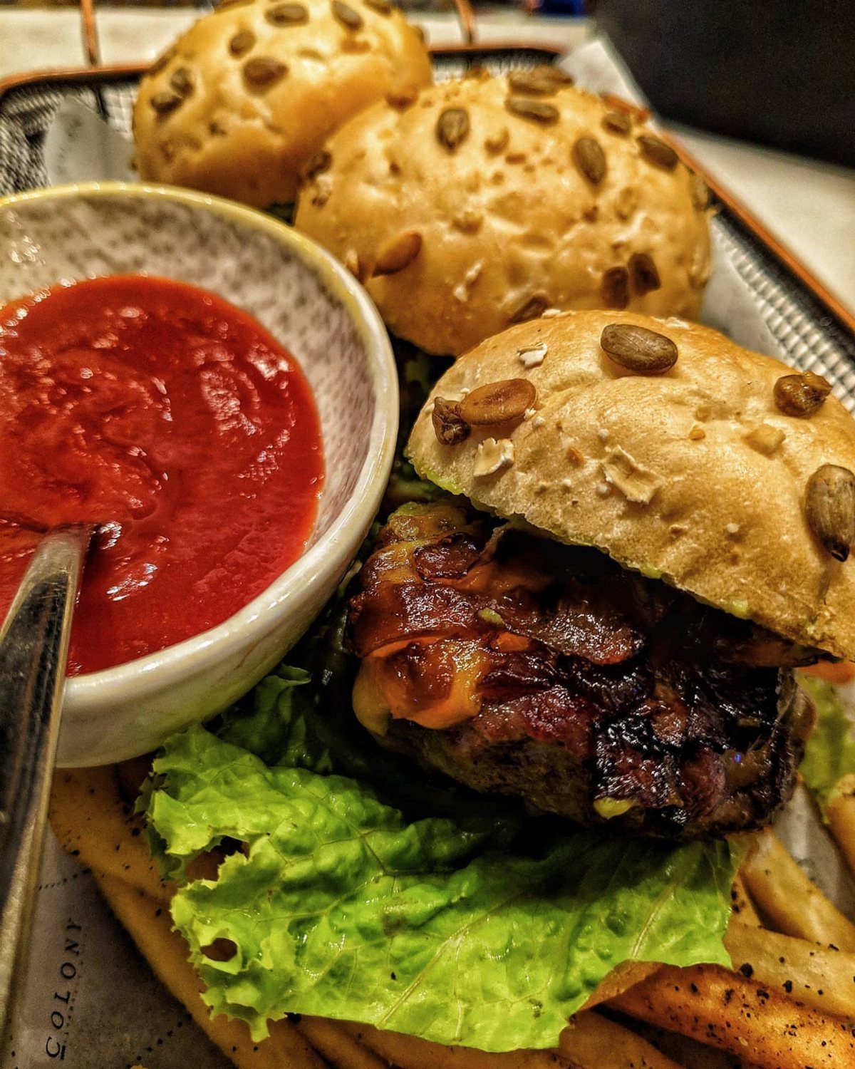 Slider with seeded bun, grilled patty, lettuce, fries, and red dipping sauce in a wire basket.