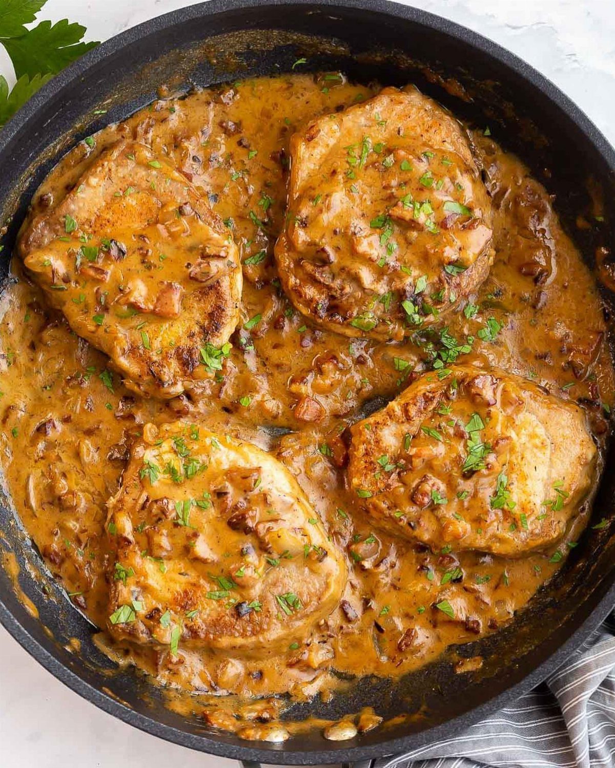 Pork chops in creamy onion sauce with parsley garnish inside a black cast-iron skillet on a marble surface.