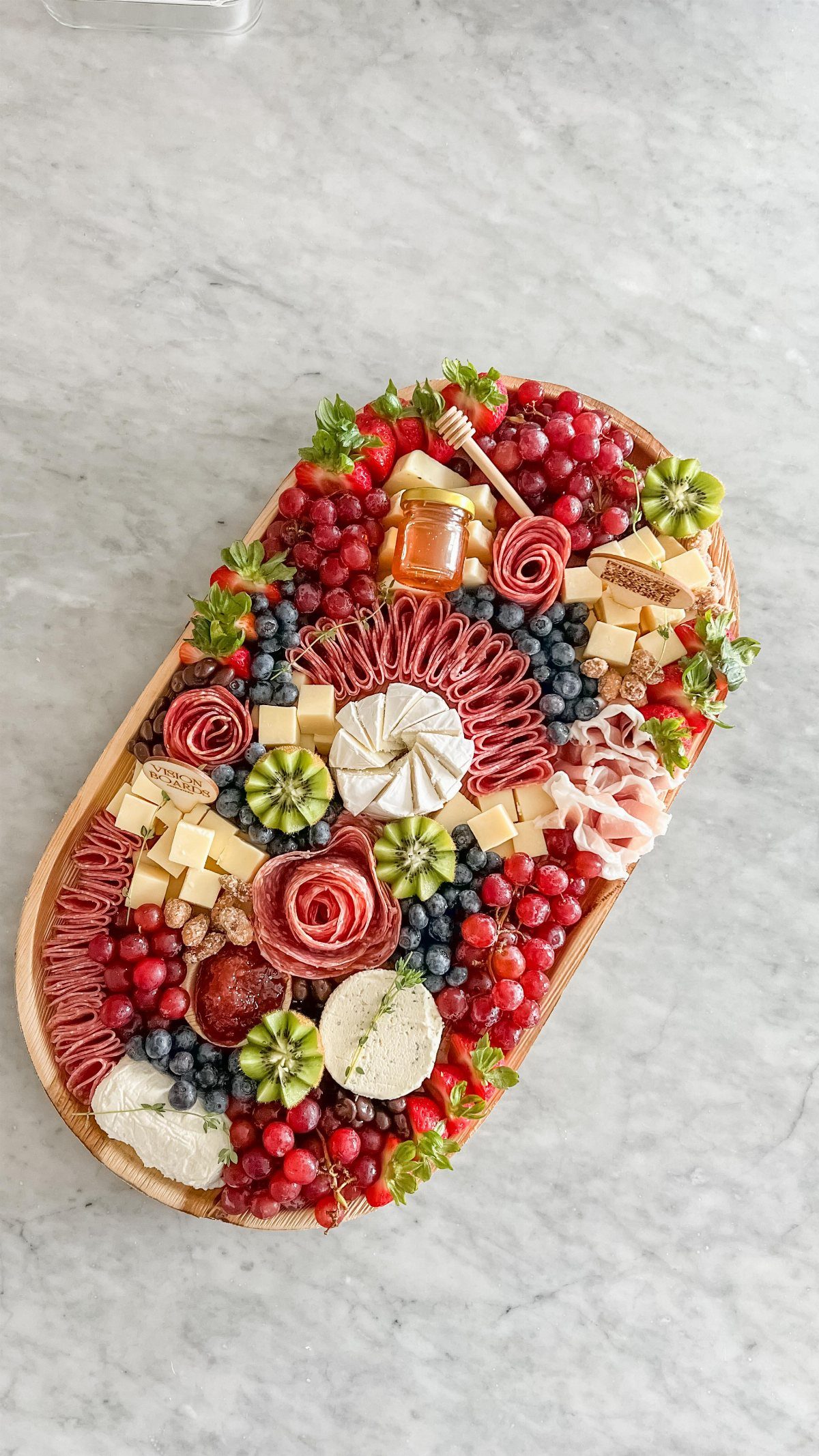 A wooden platter overflowing with assorted meats, cheeses, berries, and kiwi slices on marble.