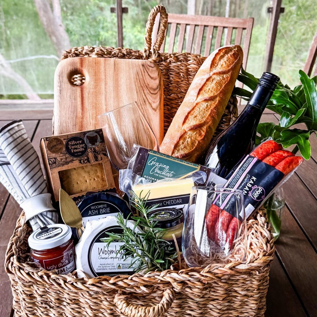 A wicker gift basket overflowing with gourmet food, wine, cheese, and a baguette on a wooden deck.