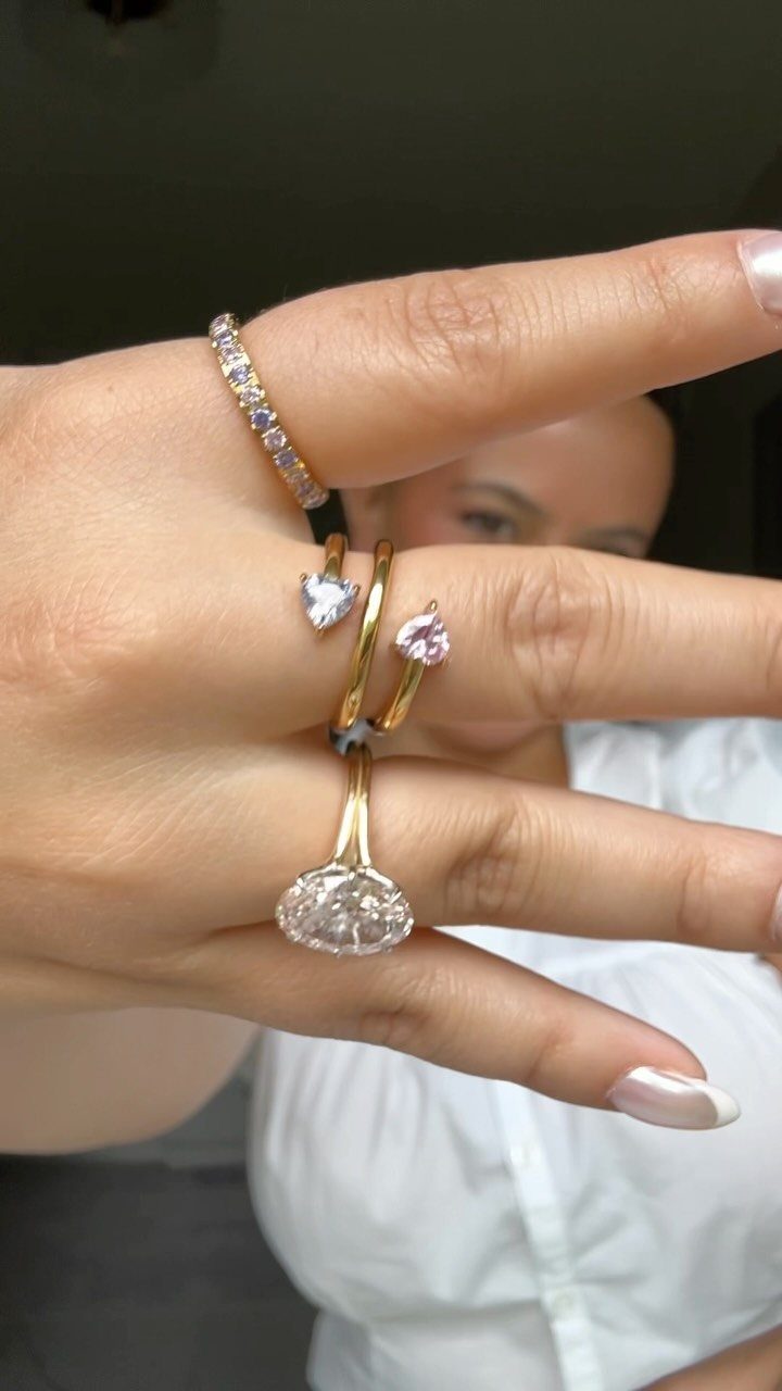 A hand wearing three gold rings with oval, heart, and multicolored stones.