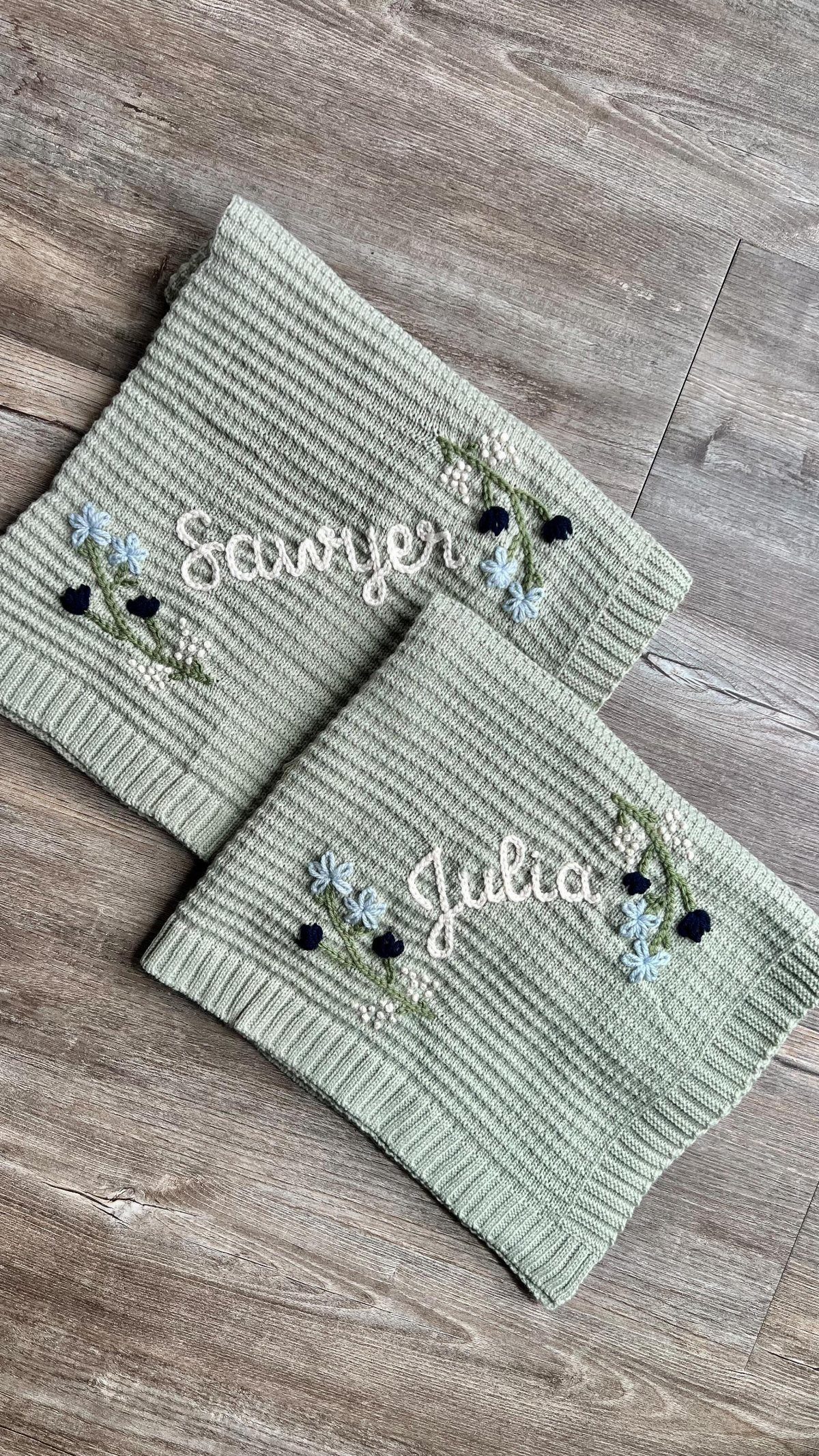 Two sage green ribbed fabric pieces with embroidered names and small floral patterns on wood.