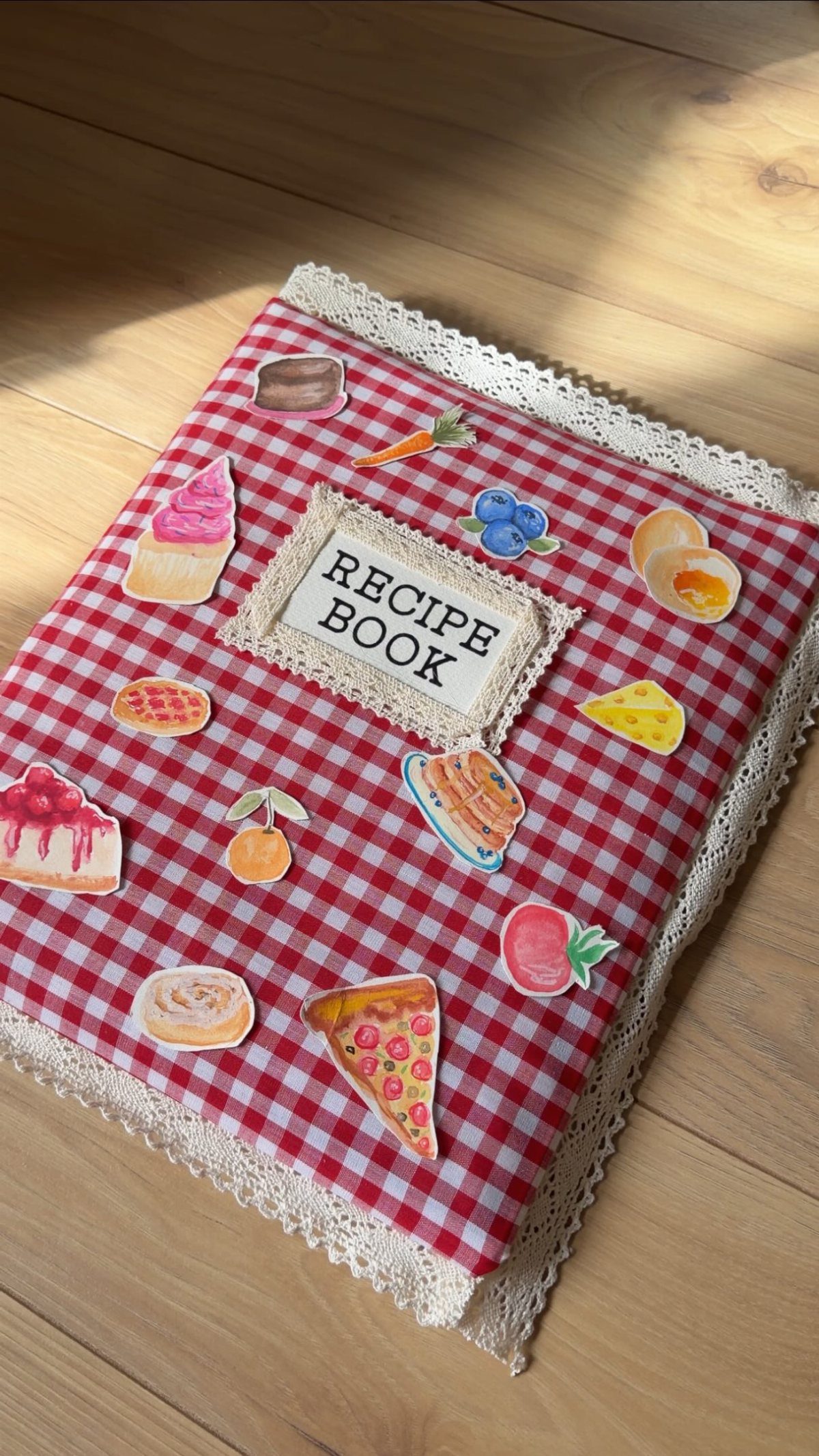 A handmade red checkered fabric recipe book decorated with colorful food illustrations and lace.