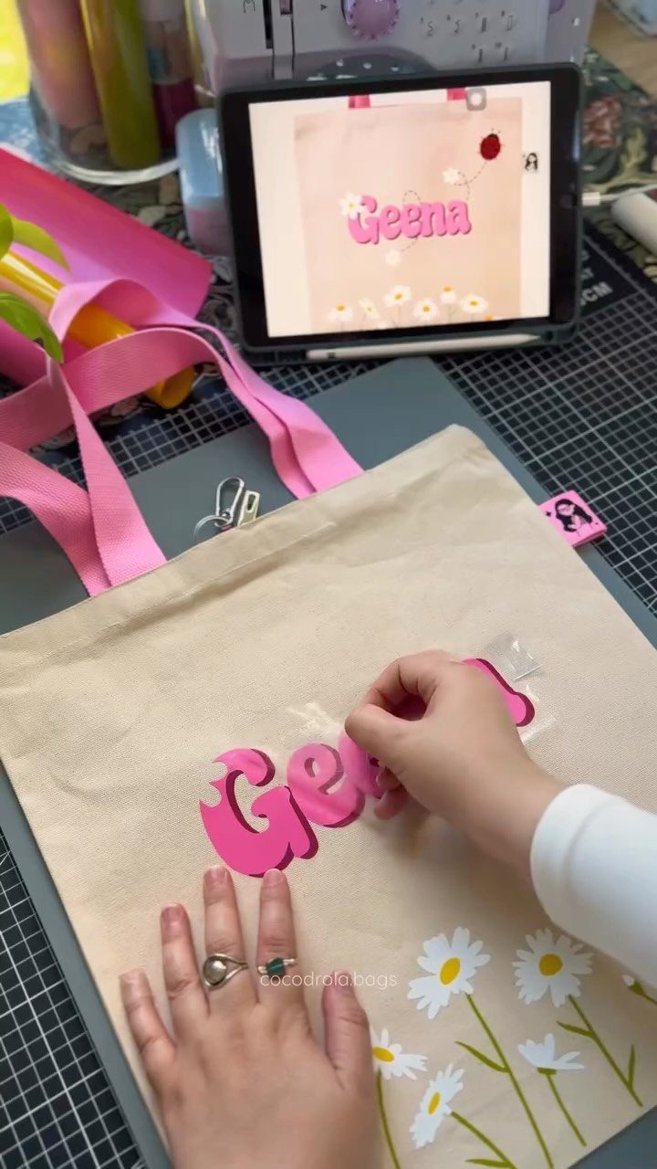 Hands applying pink letters and daisy decals onto a beige canvas tote bag.