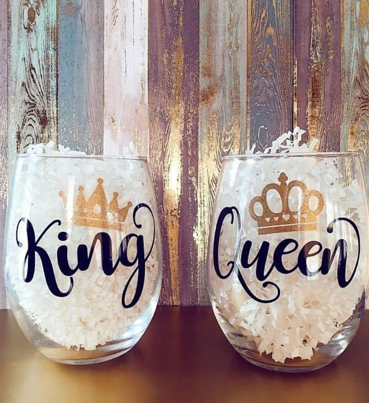 Two stemless wine glasses filled with white paper, featuring gold crowns and black text reading King and Queen.