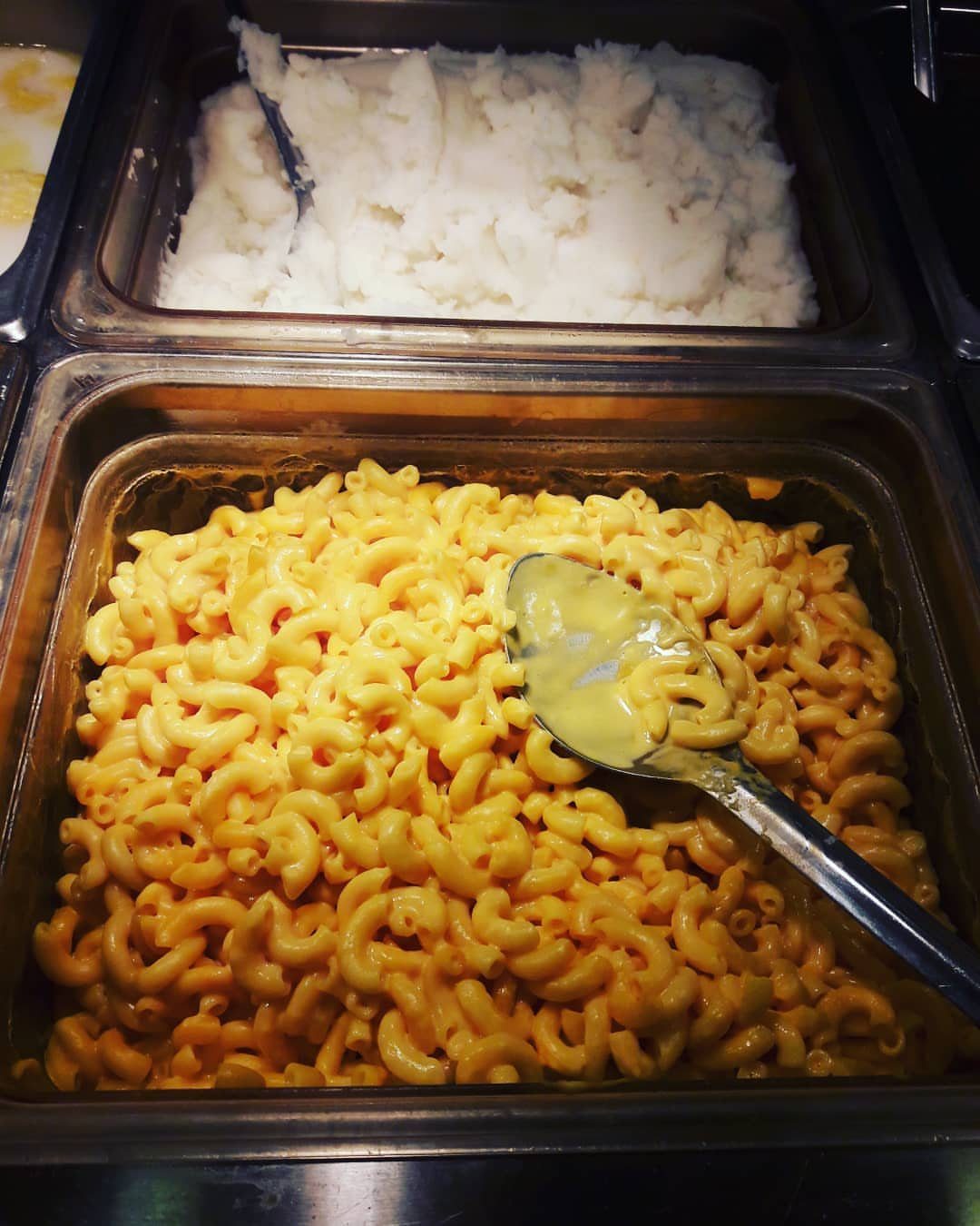 Stainless steel buffet trays holding macaroni and cheese with a spoon and a separate tray of white mashed potatoes.