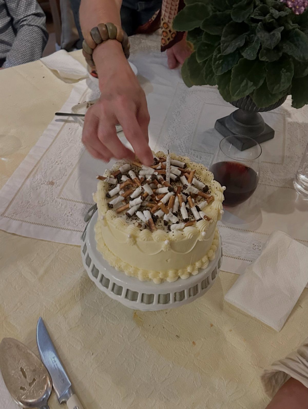 Hand reaching for small white cake decorated with cigarette butts and one candle on a table with wine and flowers.