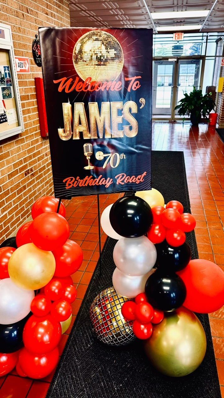 A black birthday welcome sign for James' 70th roast surrounded by red, gold, and black balloons.