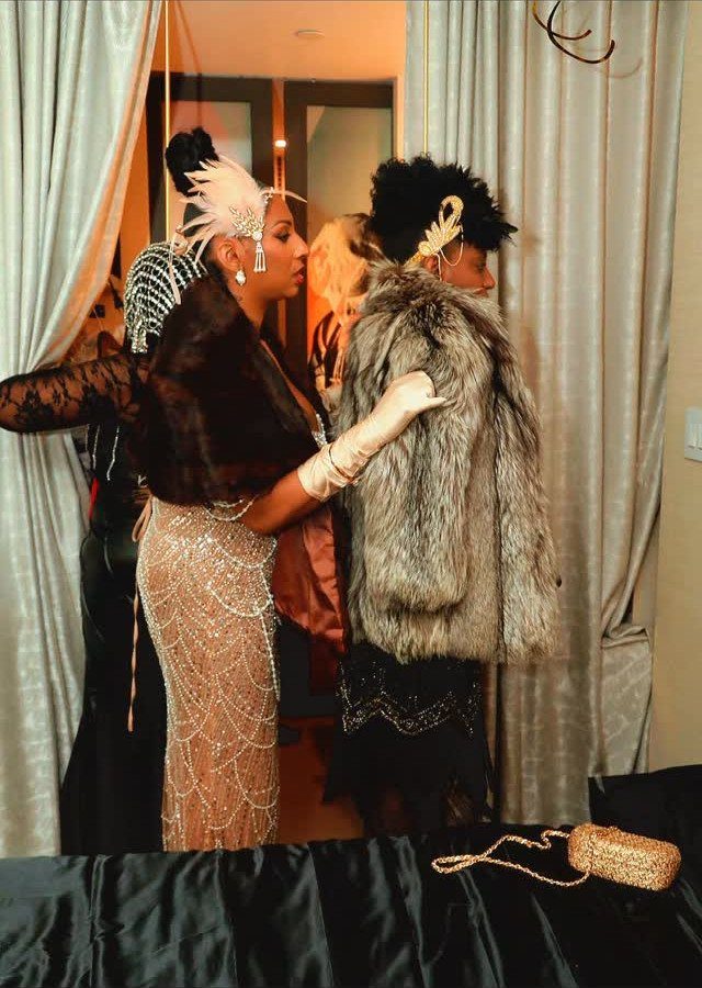 Two women in vintage flapper costumes with fur coats and feathered headbands standing indoors.