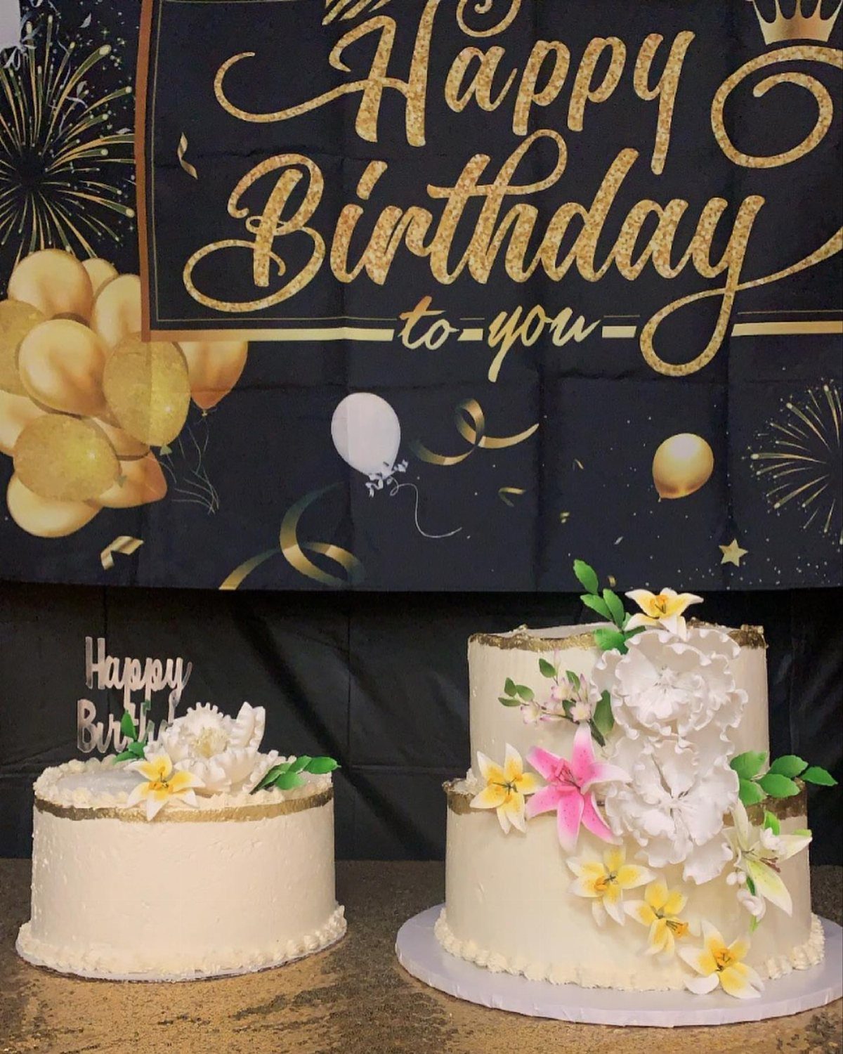 Two white floral cakes with gold trim displayed in front of a black and gold Happy Birthday banner featuring balloon graphics.