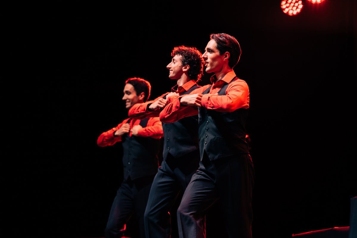 Three male performers in orange shirts and black vests dancing on a dark stage.
