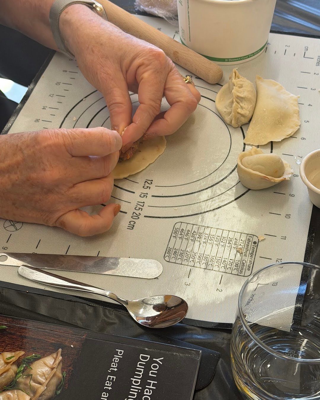 Elderly hands folding dumplings on a silicone mat with a rolling pin, spoons, and a printed card.