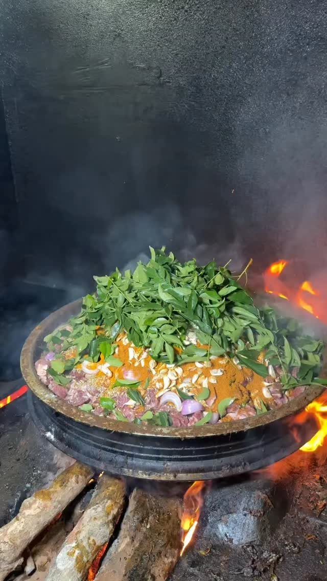 Large metal pot cooking over open wood fire filled with spiced meat, onions, and fresh green leaves.