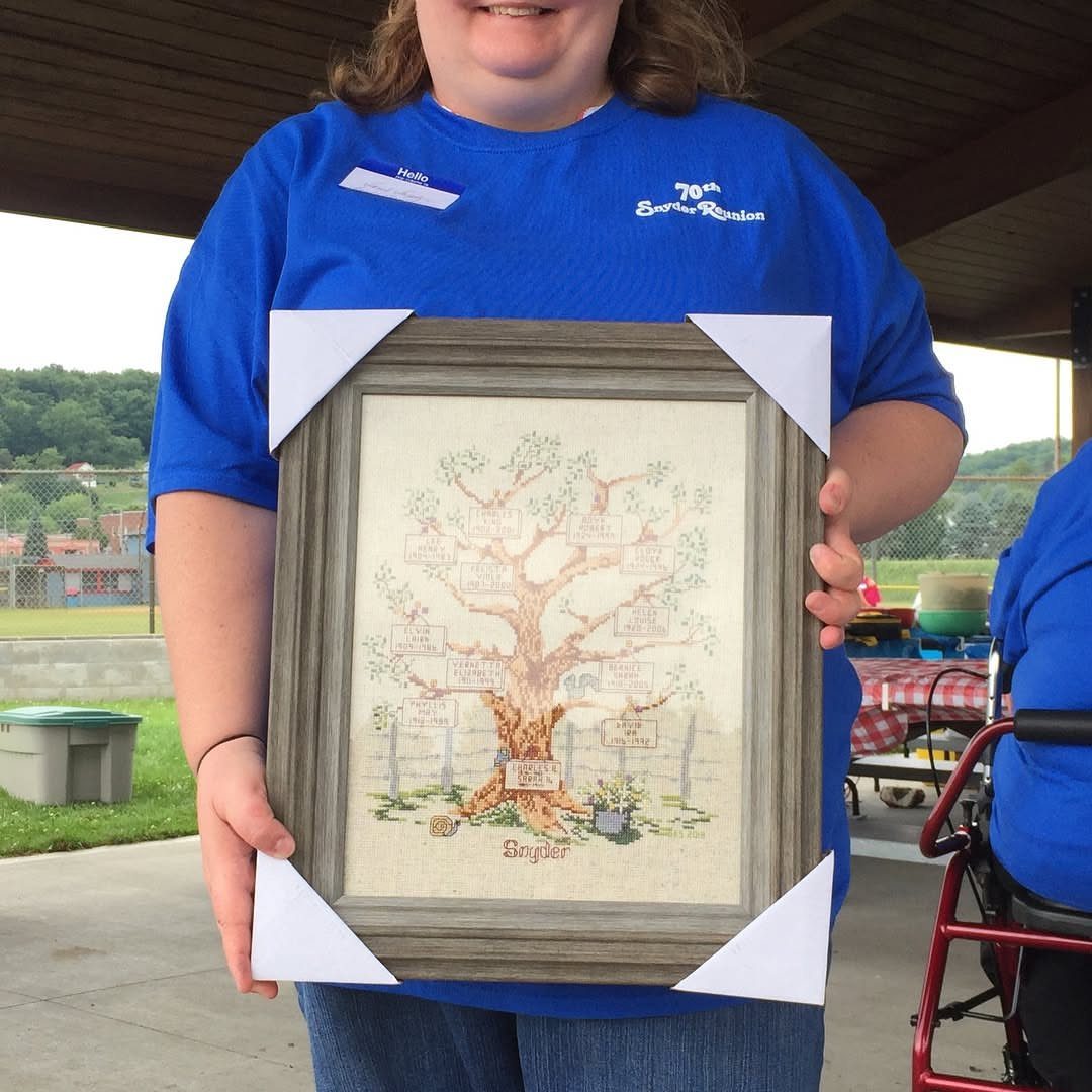 Person holding framed family tree art at outdoor reunion wearing blue shirt with 70th birthday text.