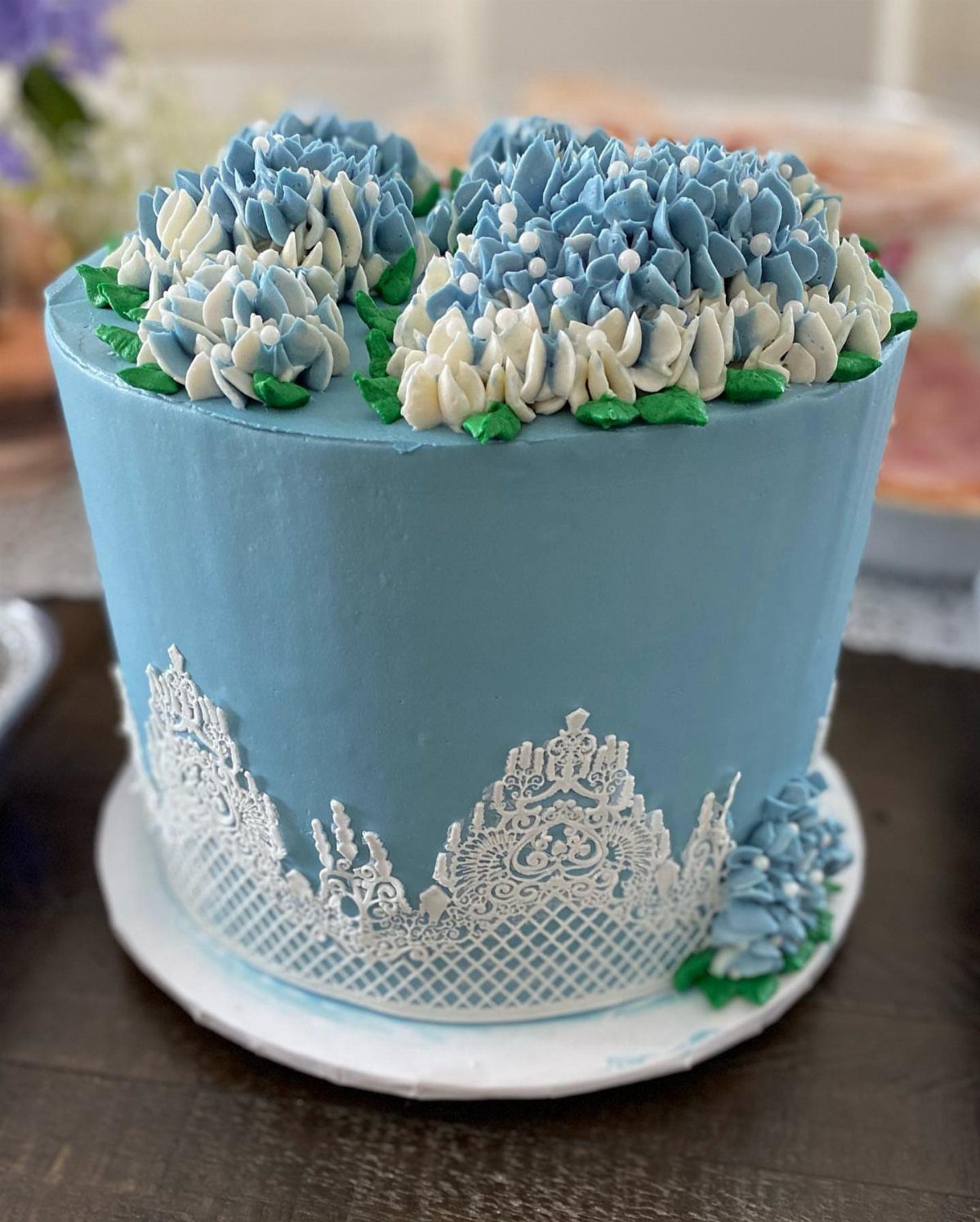 Light blue frosted cake with white lace icing and blue and white buttercream flowers on top.