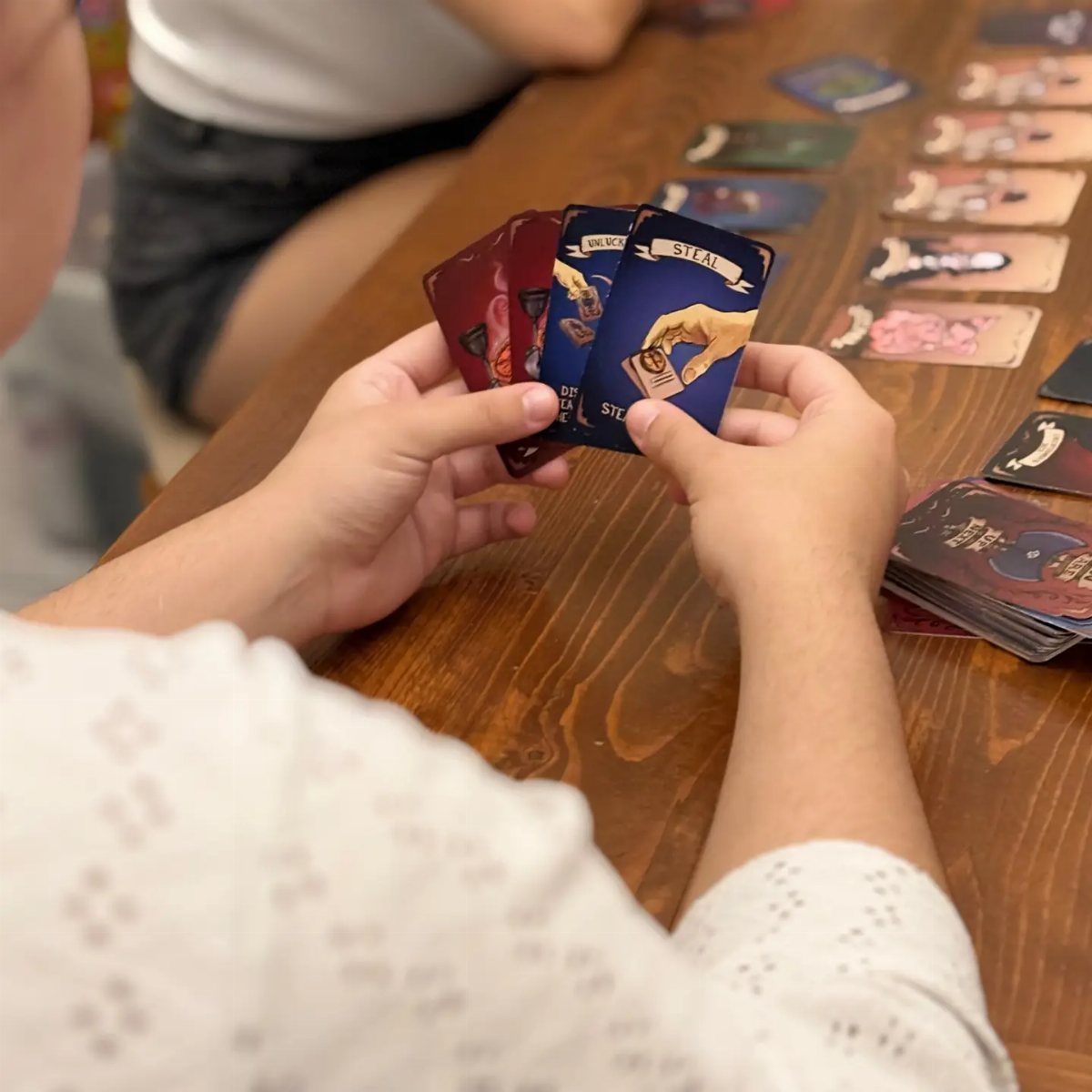 Hands holding illustrated game cards over a wooden table where other cards are arranged in rows for play.