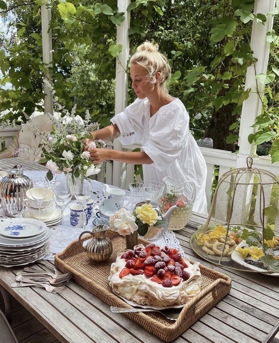 Blonde woman arranging flowers on an outdoor garden table set with blue china, strawberry meringue cake, and pastries.