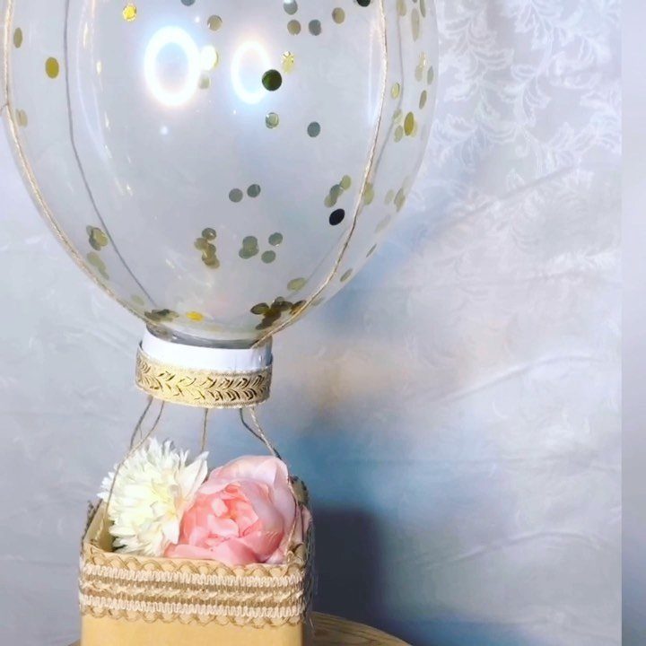 A clear balloon with gold confetti floating above a basket of pink and white flowers.