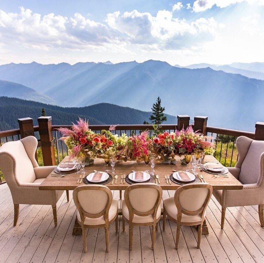 Elegant outdoor dinner table set for six on a mountain deck with pink floral arrangements and scenic views.