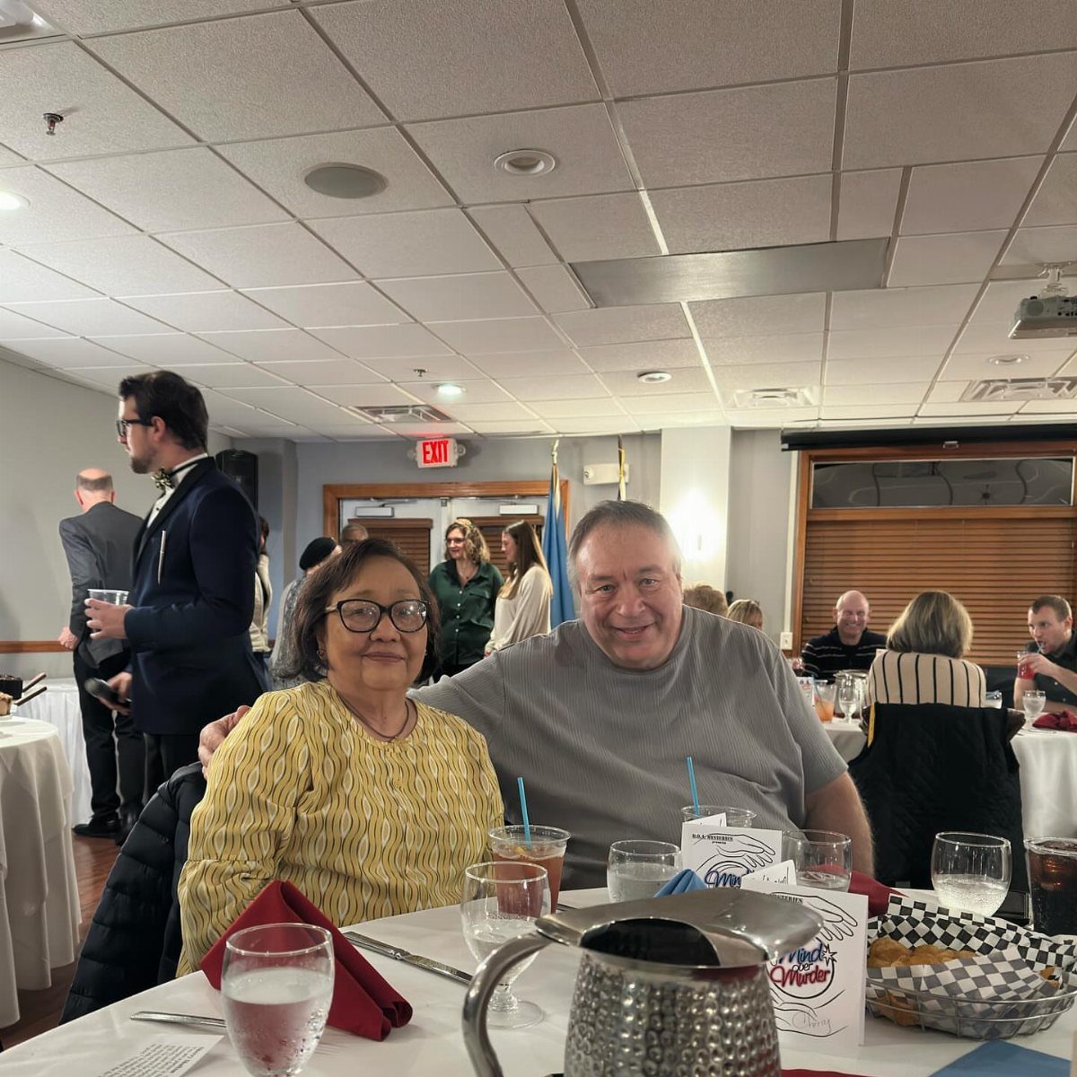 Smiling couple at a banquet table with Murder Mystery dinner menus and a man in a tuxedo standing nearby.