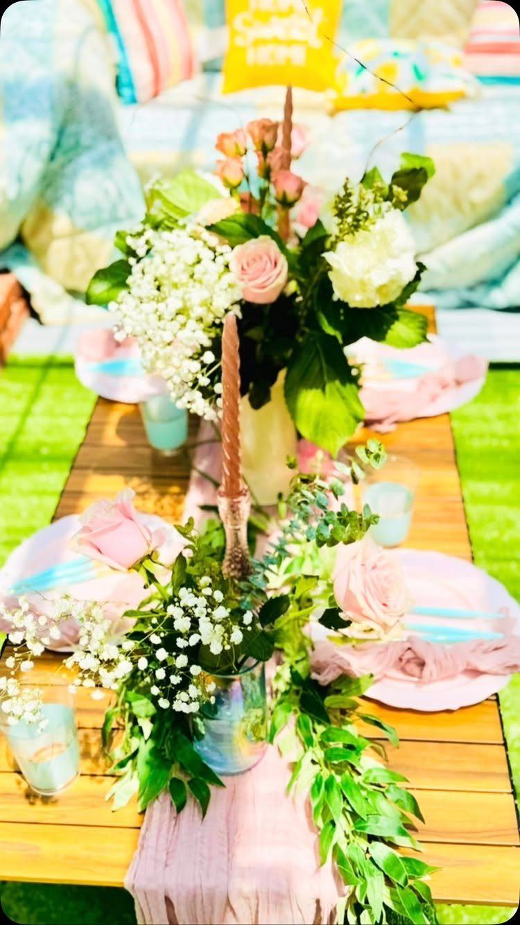 A wooden outdoor table decorated with pink plates, blue glasses, and white and pink floral arrangements.