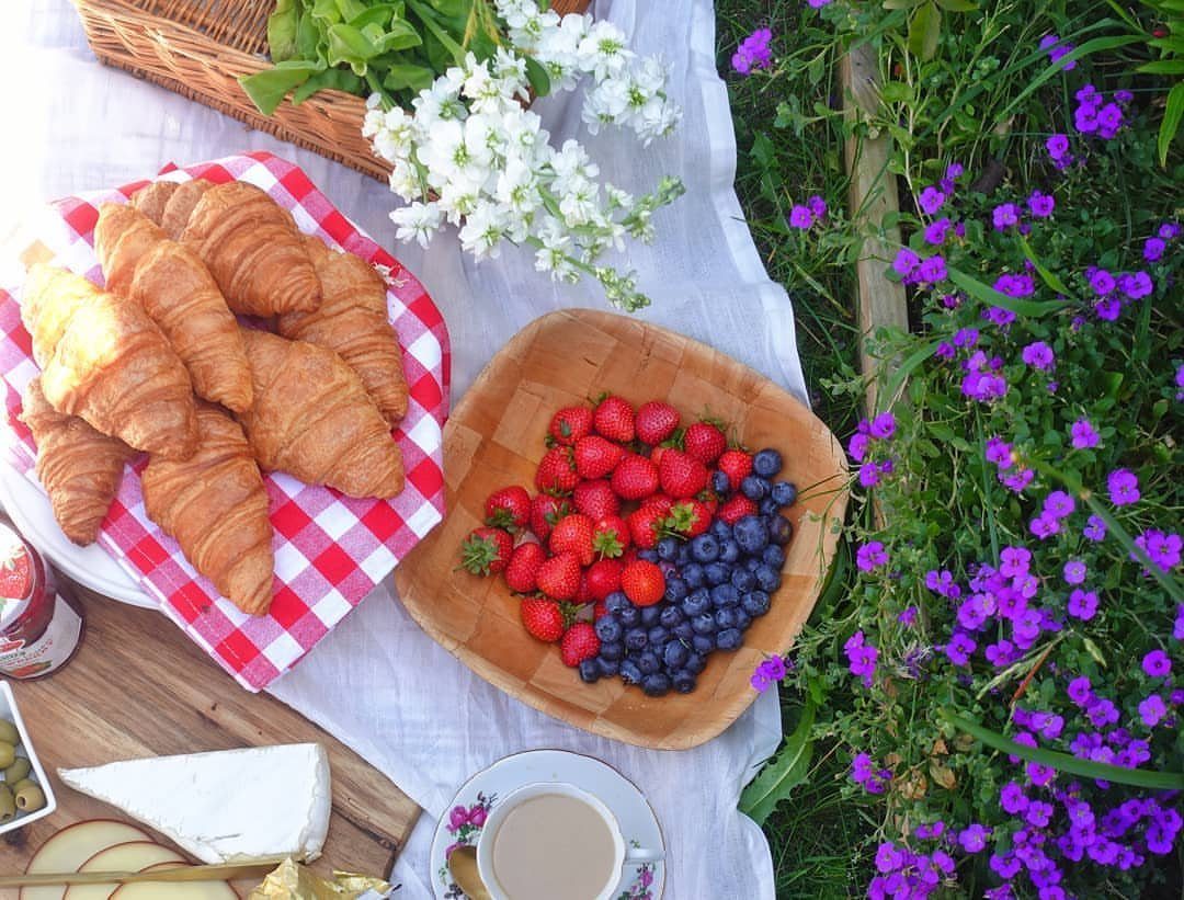 Picnic spread with croissants, berries, cheese, and tea on a white blanket next to purple garden flowers.