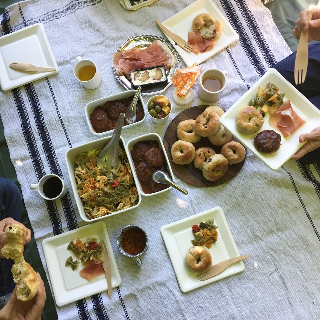 Overhead shot of a potluck picnic spread with bagels, meats, and salads on a striped blanket with people serving themselves.