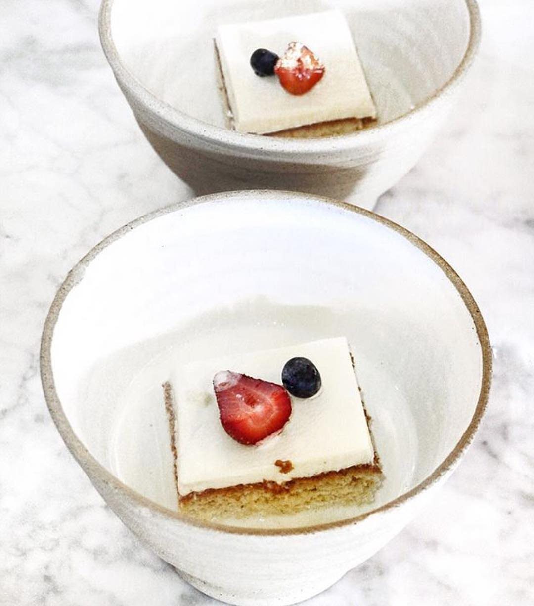Two elegant individual dessert squares topped with strawberries and blueberries served in rustic bowls on a marble table.