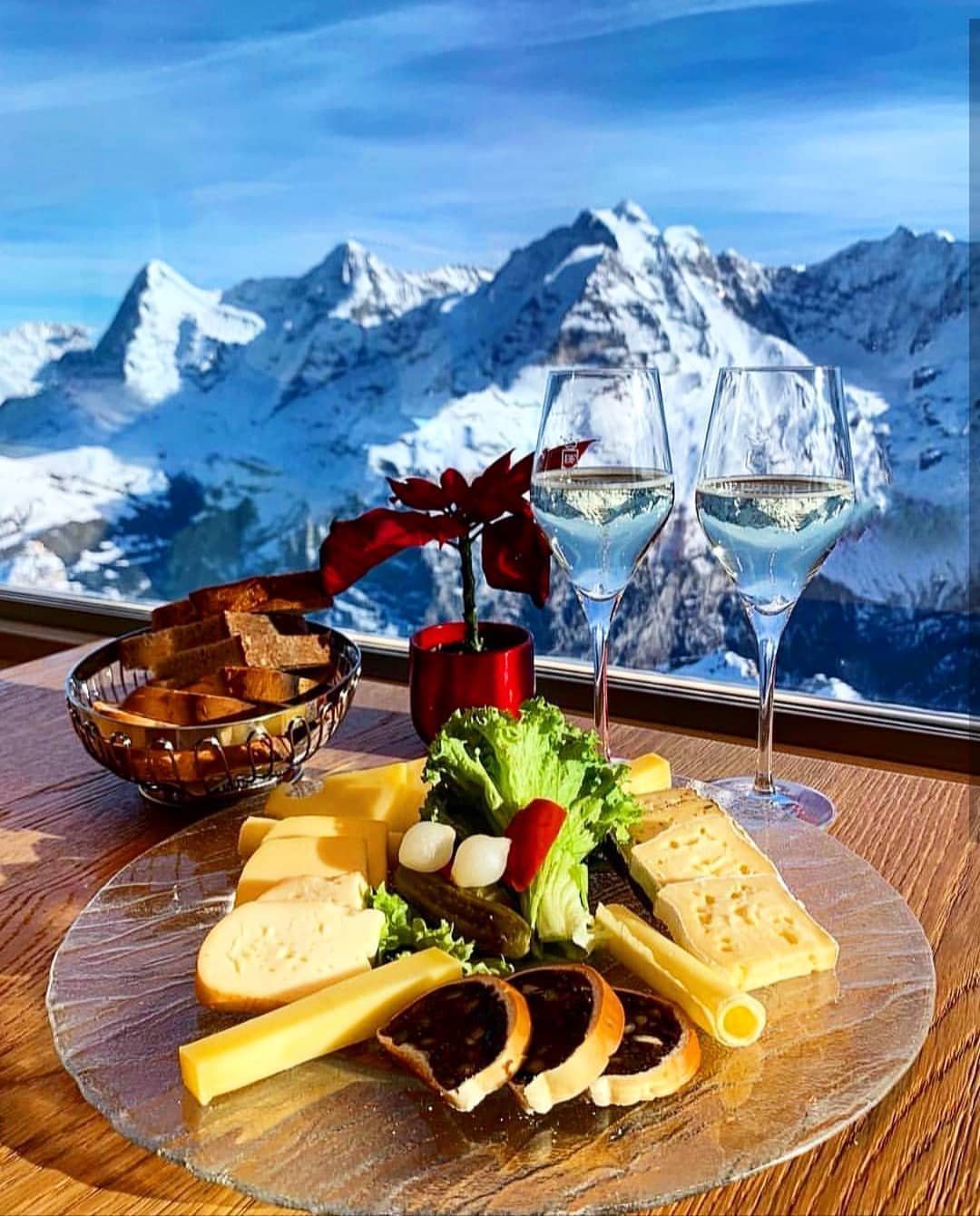 Two glasses of white wine and a cheese platter on a table with a snowy mountain view through the window.