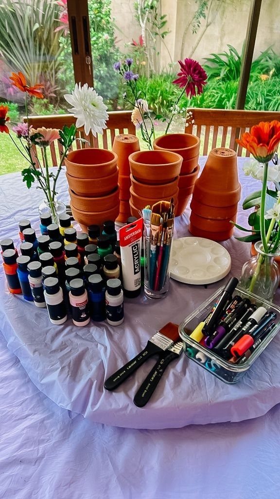 Terracotta pots, acrylic paint bottles, and various brushes arranged on a purple tablecloth outdoors.