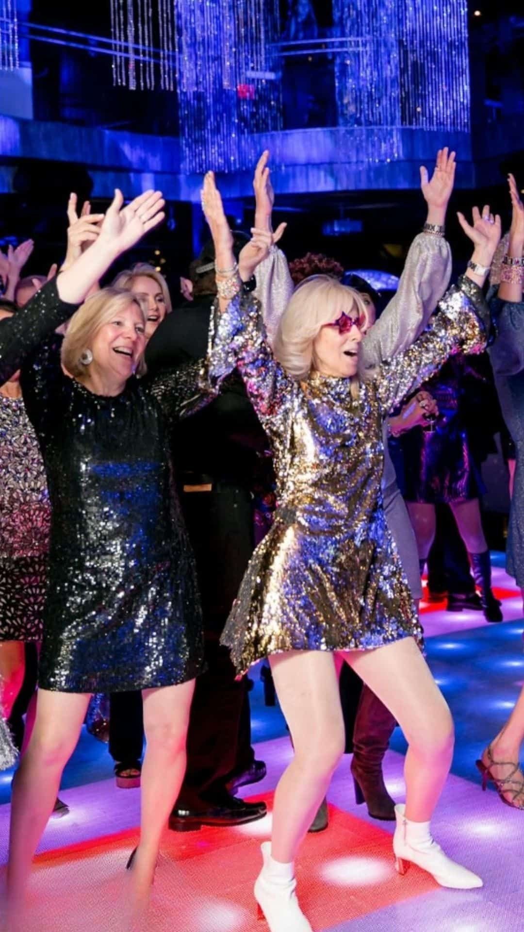 Two women in sparkling sequined dresses dancing joyfully with raised arms under a crystal chandelier.