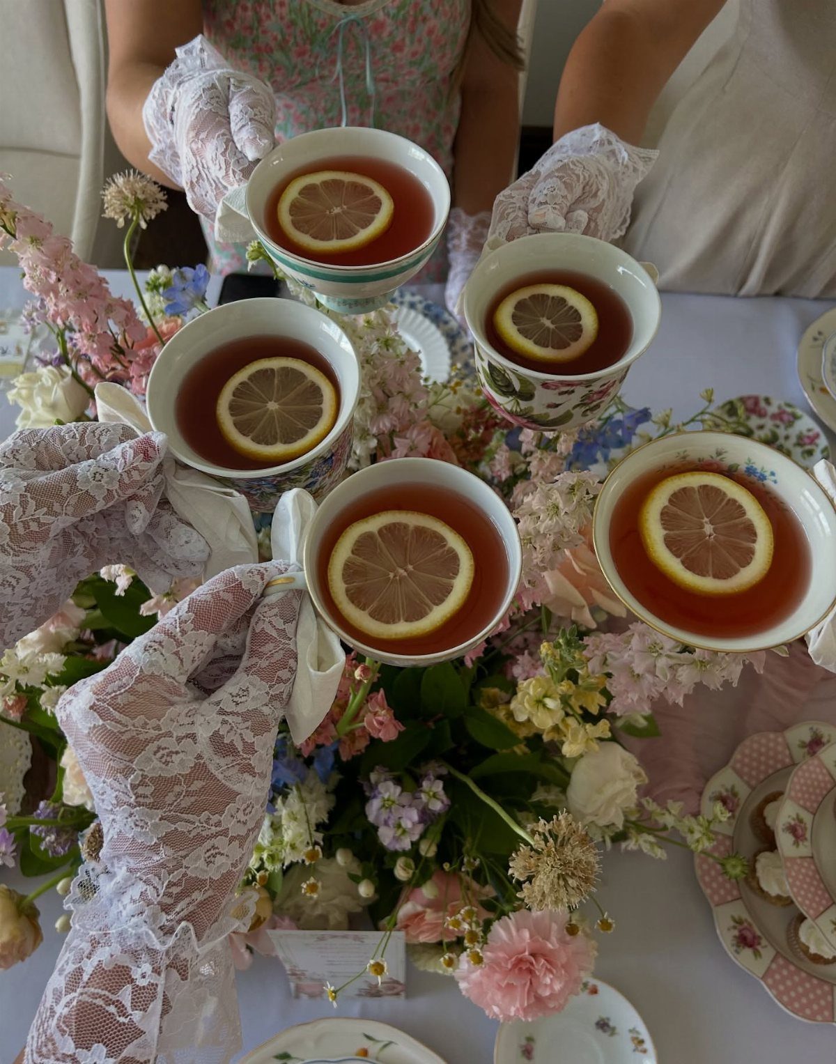 Hands in lace gloves holding teacups with lemon slices over a floral table setting.
