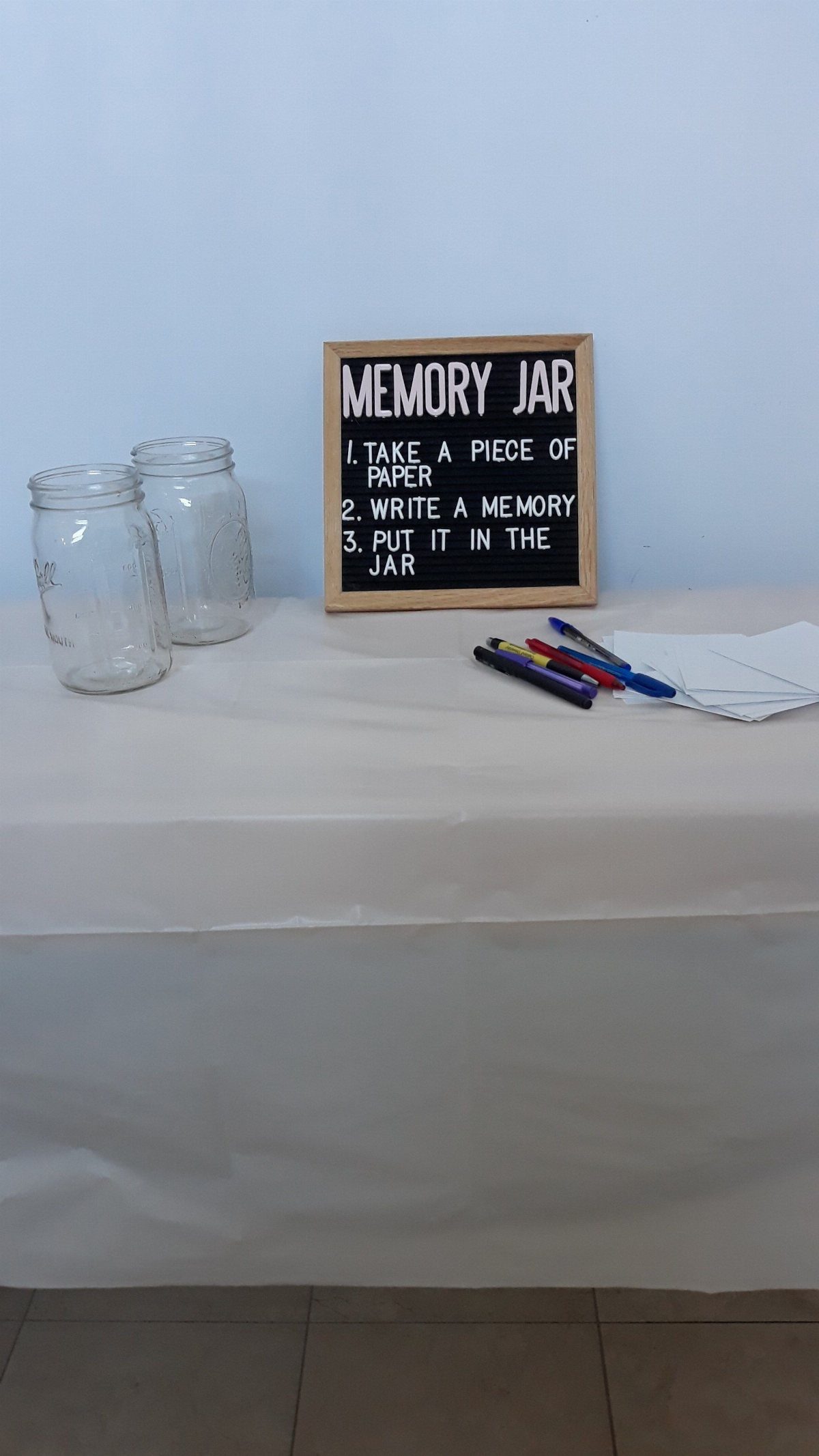 Two glass jars and a memory jar sign with pens and paper on a white table.