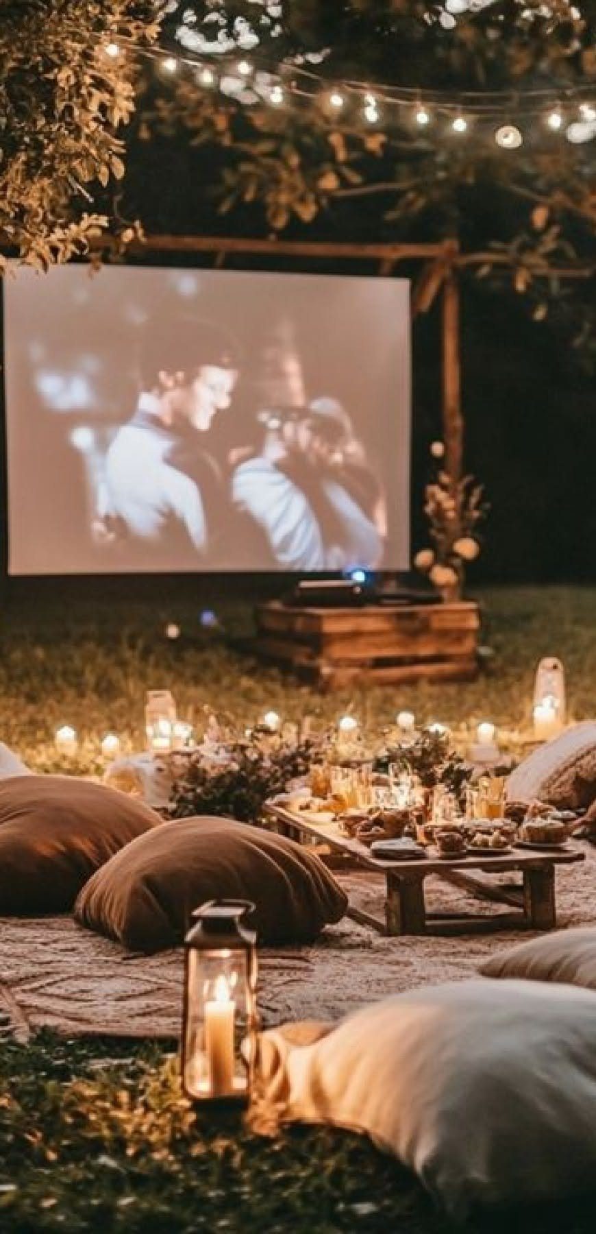 Outdoor movie night setup with a projection screen, lanterns, candles, and floor cushions on grass.