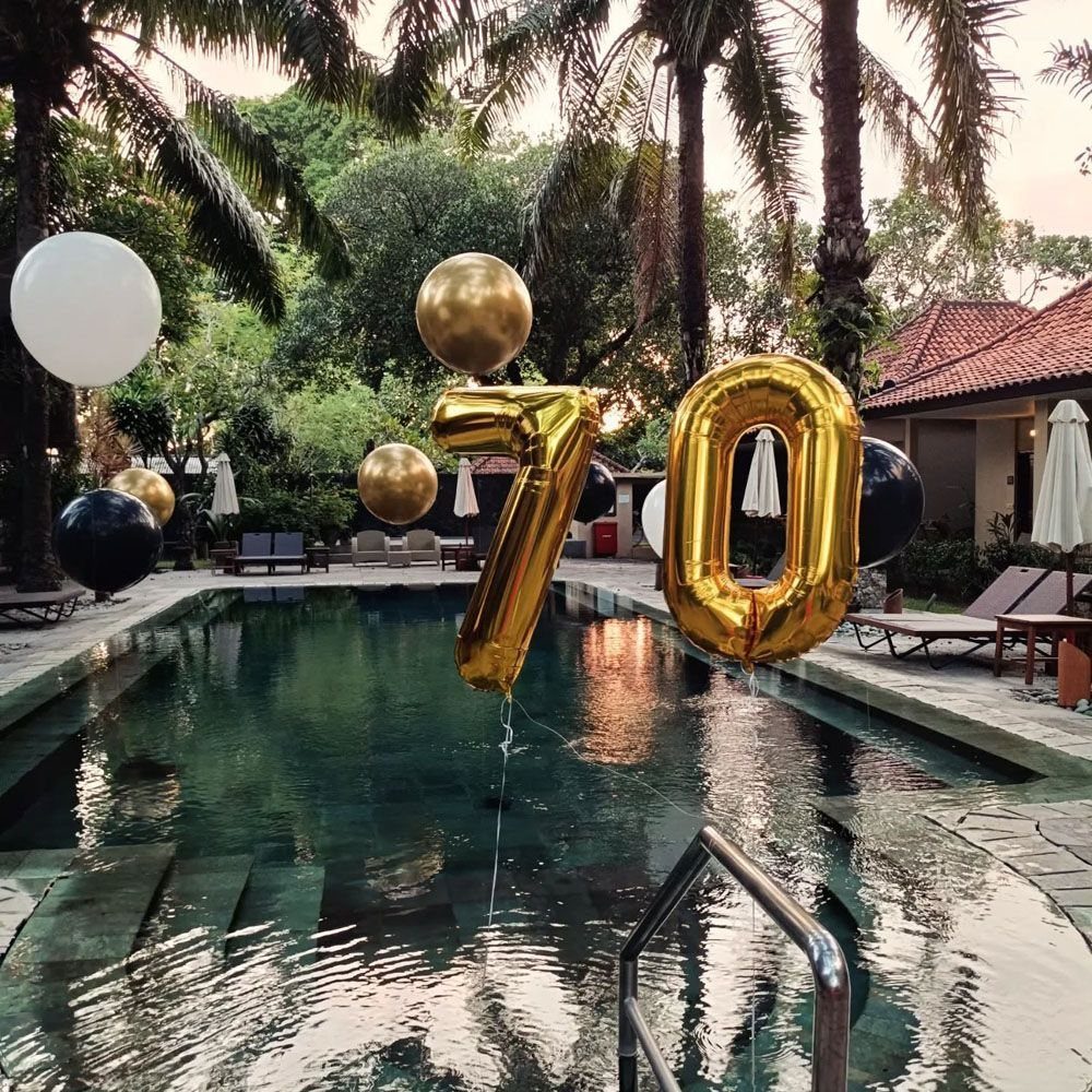 Gold number seventy balloons floating over a swimming pool surrounded by palm trees.