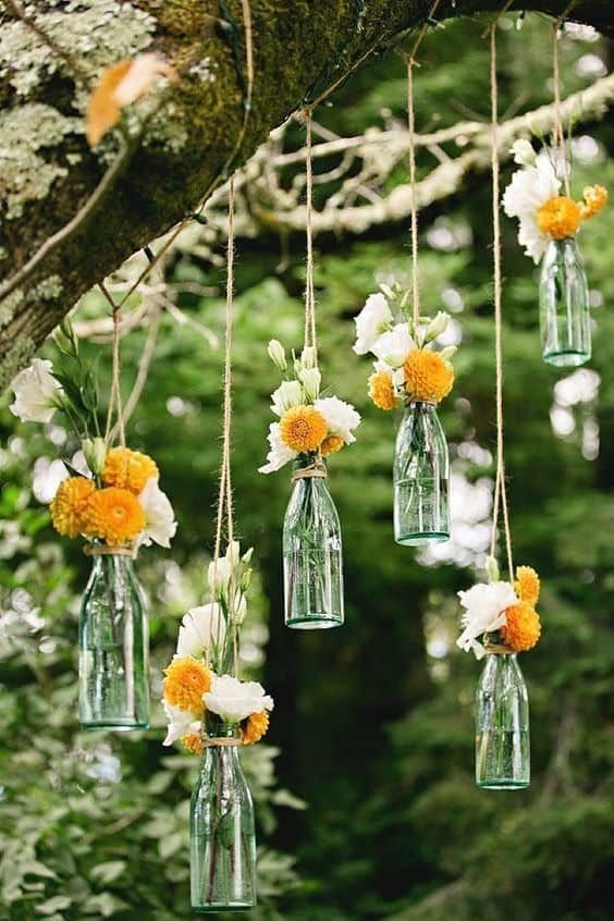 Green glass bottles with white and orange flowers hanging from a tree branch by twine.