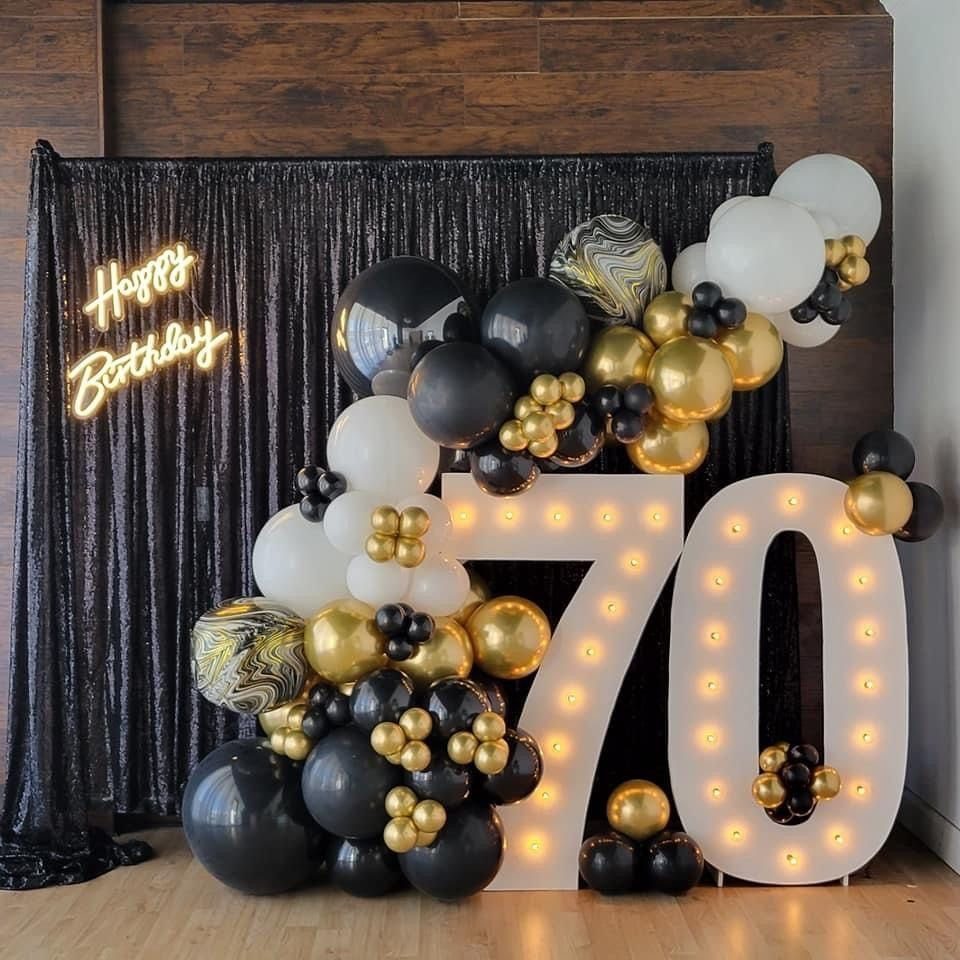 Large illuminated number 70 surrounded by black, white, and gold balloons against a dark curtain.
